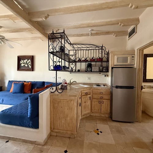 Un espacio interior que presenta una sala de estar con un sofá azul, una cocina integrada de estilo rústico con electrodomésticos modernos, y un techo con vigas de madera.