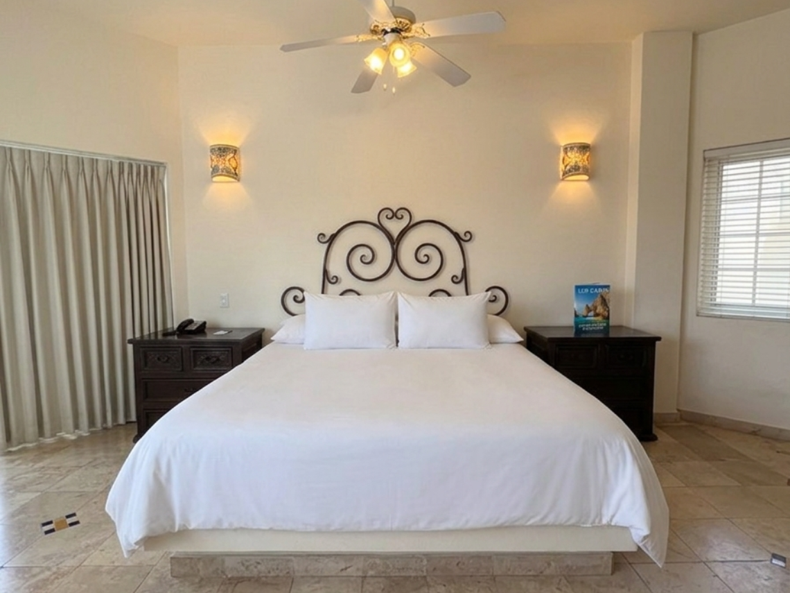 A light-filled bedroom features a large bed with an ornate headboard and red petals, a ceiling fan, patterned tiled flooring, and a sliding glass door leading to an outdoor patio with a view of cars and vegetation.