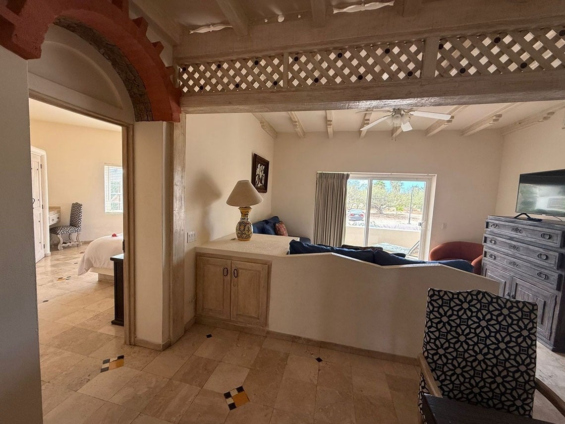 Una vista de un interior hogareño con un arco que conduce a un dormitorio, y un área de sala de estar con sofá, televisor y aparador, todo bajo un techo de vigas y un enrejado decorativo.