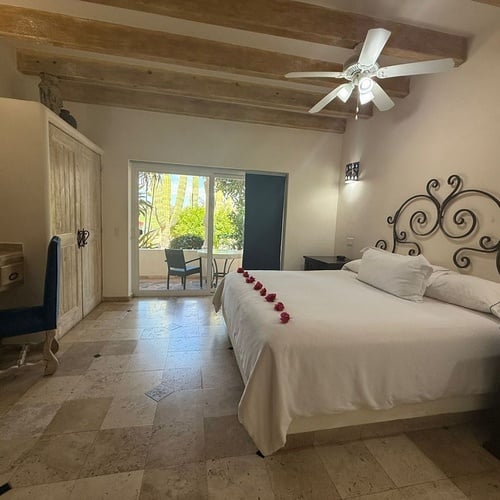 Un dormitorio de estilo rústico presenta una cama adornada con pétalos de rosa, mobiliario de madera, techo con vigas expuestas, suelo de baldosas y acceso a un balcón con vegetación.