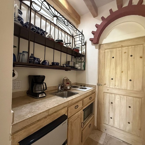 Una cocineta de estilo rústico con electrodomésticos básicos y estantes de hierro forjado llenos de vajilla, junto a una puerta de madera bajo un arco decorativo.