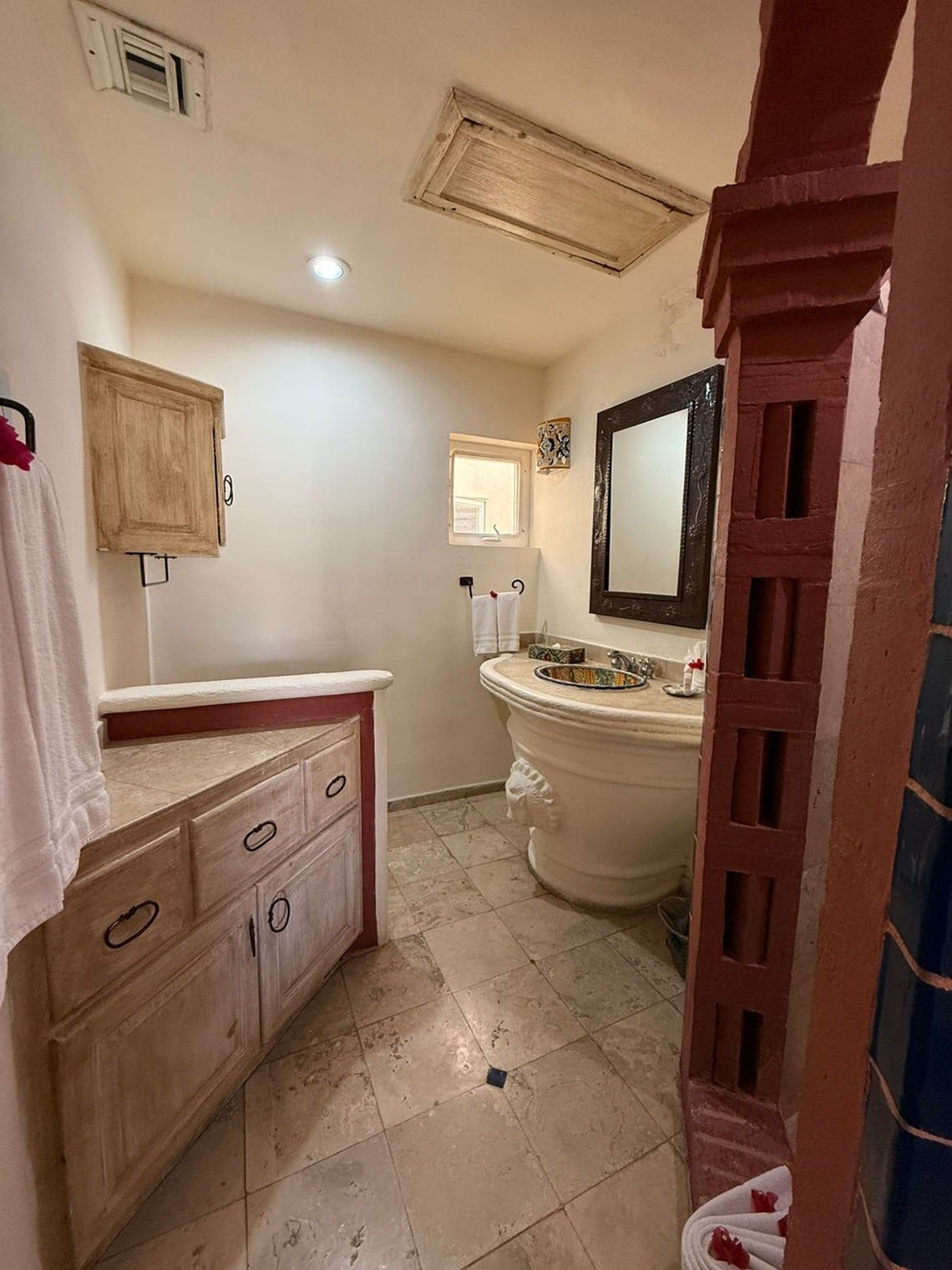 Un baño de estilo rústico y tonos cálidos, con un lavabo pedestre ornamentado, un mueble de esquina de madera clara y un suelo de baldosas.