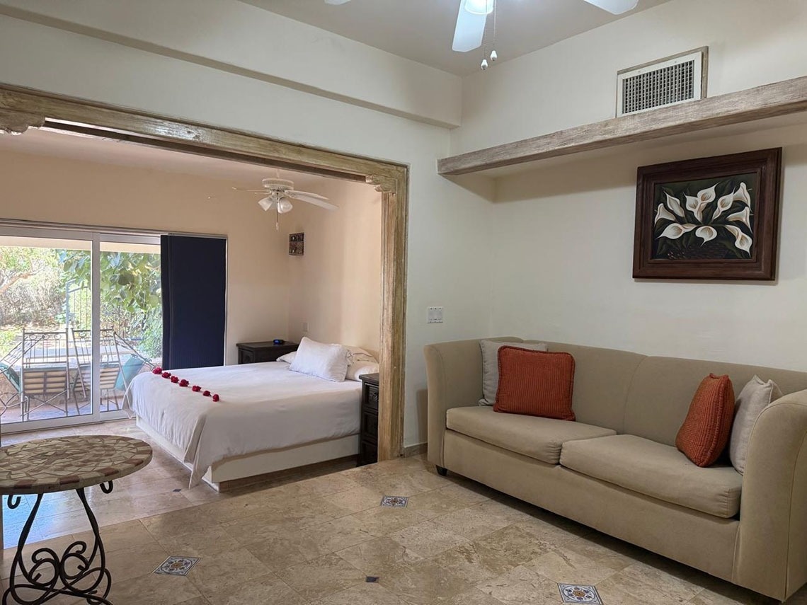 Una habitación interior fusiona un dormitorio con una cama blanca decorada con pétalos rojos y una sala de estar con un sofá beige, ambos con suelo de baldosas y acceso a un patio exterior a través de puertas corredizas.