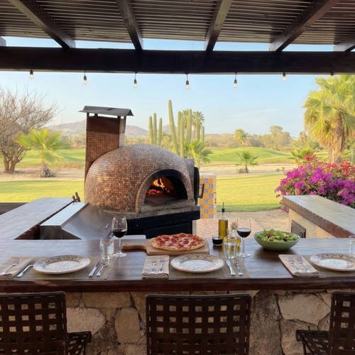 An outdoor patio shaded by a large fabric canopy with string lights features a stone-clad bar with a pizza oven, multiple seating areas, and overlooks a golf course with desert vegetation and mountains under a partly cloudy sky.