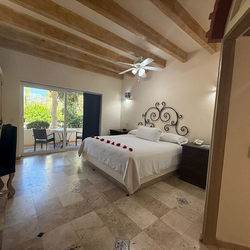 Un dormitorio de estilo rústico con vigas de madera, cama decorada con pétalos de rosa, suelo de baldosas y una puerta corredera que da a un patio exterior.