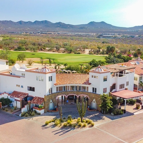 Un resort de golf en Los Cabos, con edificios blancos y techos de teja, se sitúa entre un campo de golf verde y un paisaje desértico montañoso bajo un cielo claro.