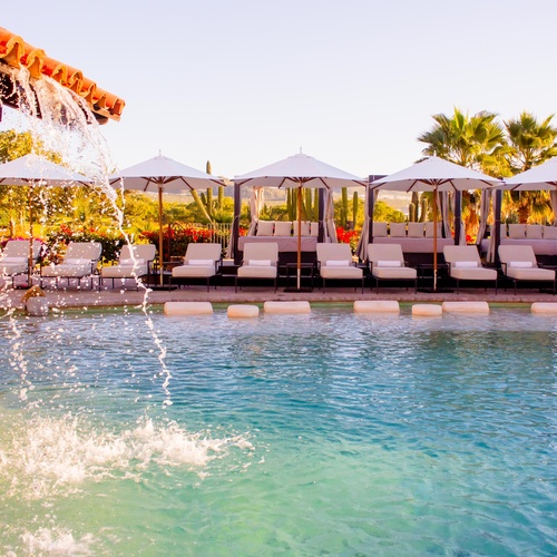 A sunny resort pool area features a swim-up bar with a red-tiled roof, surrounded by Mediterranean-style buildings, lounge chairs, and outdoor dining spaces.