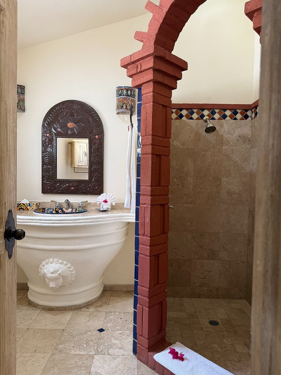 Se muestra un baño rústico con un distintivo arco de ladrillo rojo que separa un lavabo decorativo con espejo de una ducha con azulejos.