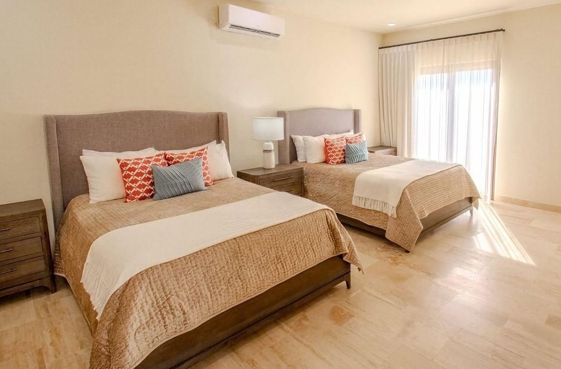 This bright bedroom features two neatly made beds with patterned pillows, a shared nightstand, an air conditioning unit, and a large window with sheer curtains allowing sunlight to illuminate the tiled floor.