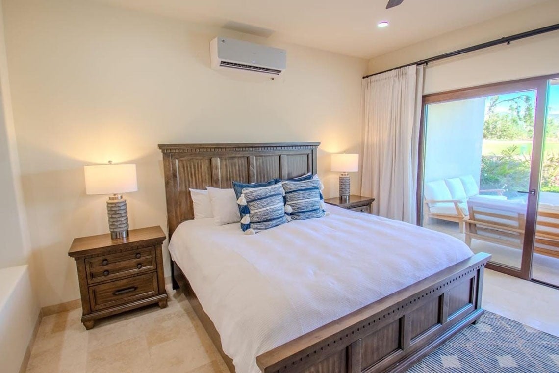 Un dormitorio bien iluminado presenta una cama grande de madera flanqueada por mesitas de noche con lámparas, un aire acondicionado montado en la pared y una puerta corrediza de vidrio que conduce a un patio exterior amueblado.