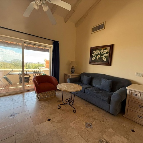 Una acogedora sala de estar con sofá, sillón y suelo de baldosas, que se abre a un balcón con vistas a un paisaje montañoso y desértico.