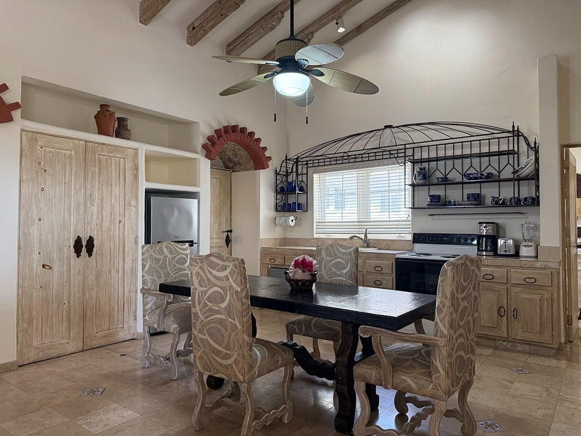 Una espaciosa cocina-comedor de estilo rústico presenta una mesa de madera, gabinetes claros, una estantería de hierro forjado sobre la ventana y un techo abovedado con vigas.