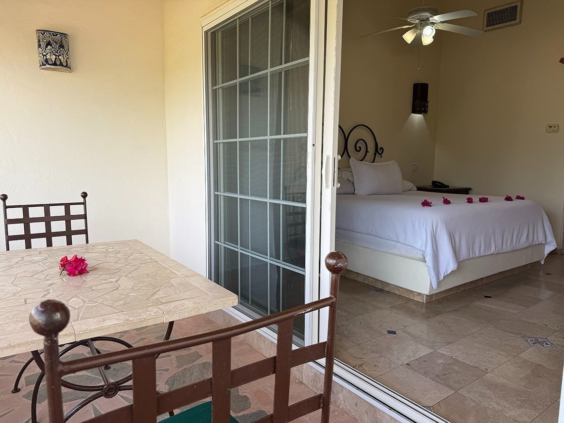 Se ve un patio exterior con mesa y silla, y a través de una puerta de cristal, un dormitorio con una cama blanca adornada con flores rojas.