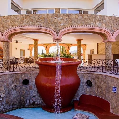 En un patio con arquitectura de piedra y arcos rosados, una gran fuente de terracota vierte agua en una piscina, rodeada de barandales de hierro forjado y áreas de descanso.