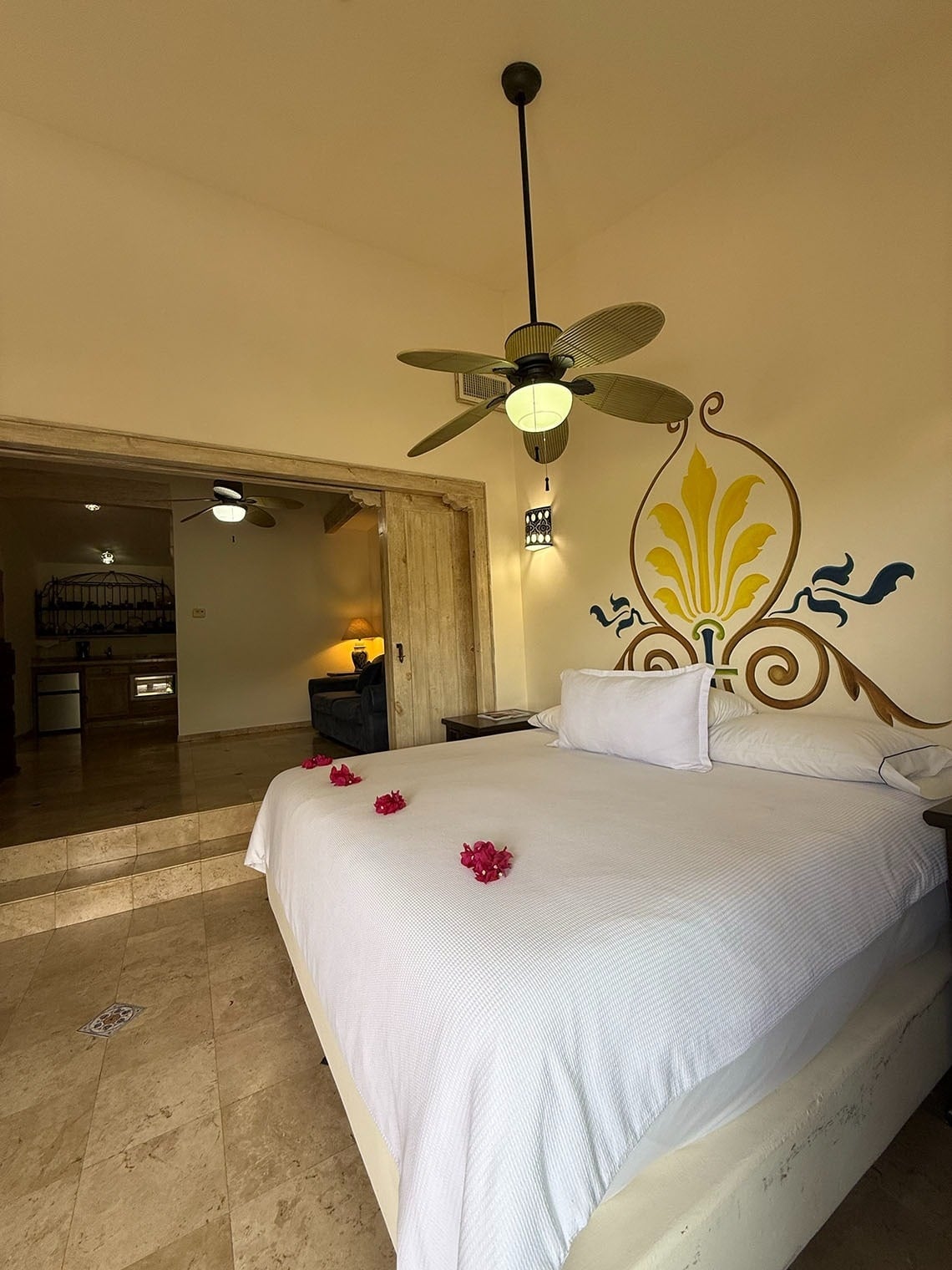 Un dormitorio cuenta con una cama decorada con pétalos rojos, un ventilador de techo, un mural ornamental en la pared y una sala contigua visible al fondo.