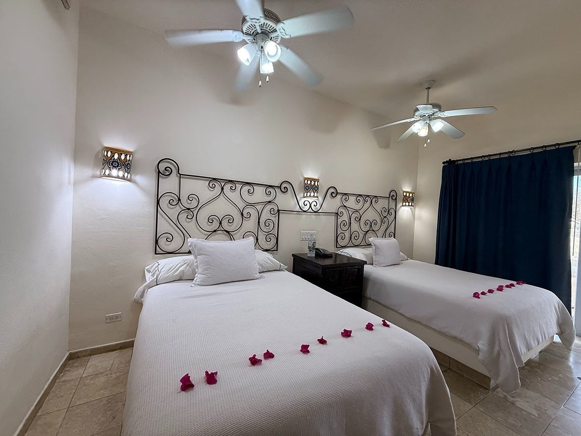 Un dormitorio presenta dos camas individuales con sábanas blancas, adornadas con pétalos rosados, cabeceras ornamentadas de hierro forjado, apliques de pared, ventiladores de techo y cortinas azules oscuras.