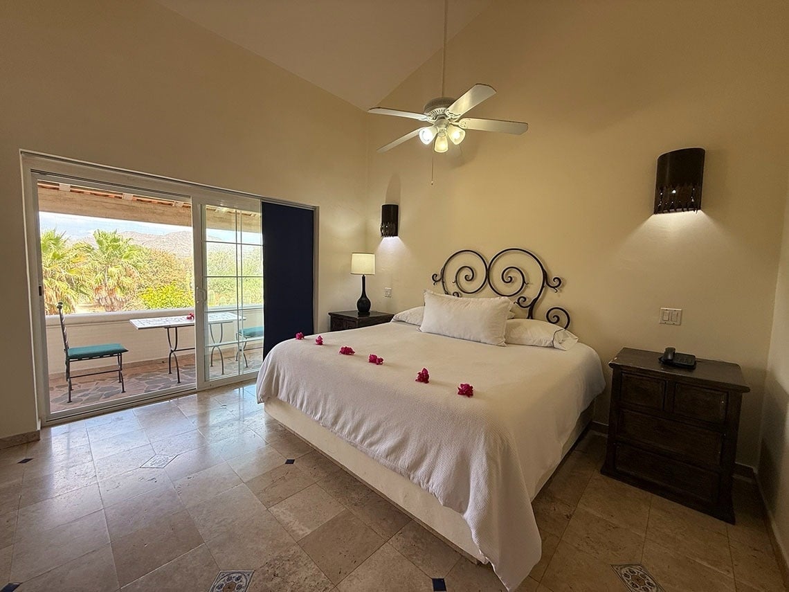 Un dormitorio luminoso con una cama grande, un ventilador de techo y un balcón que ofrece vistas a palmeras y montañas.