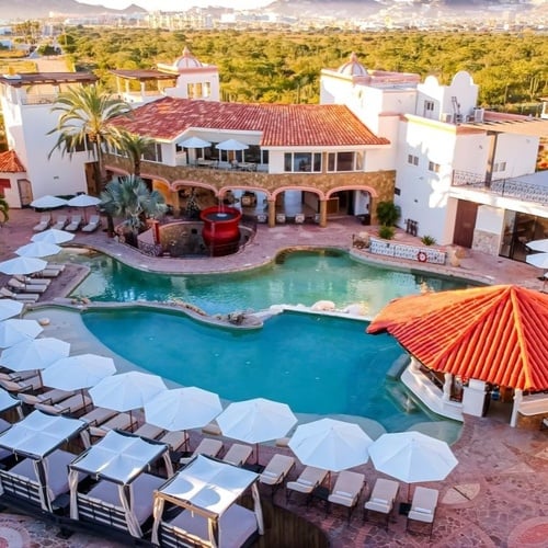 Una vista aérea muestra un lujoso complejo turístico con múltiples piscinas, tumbonas con sombrillas y cabañas, rodeado de edificios de estilo mediterráneo y vegetación, con montañas al fondo.