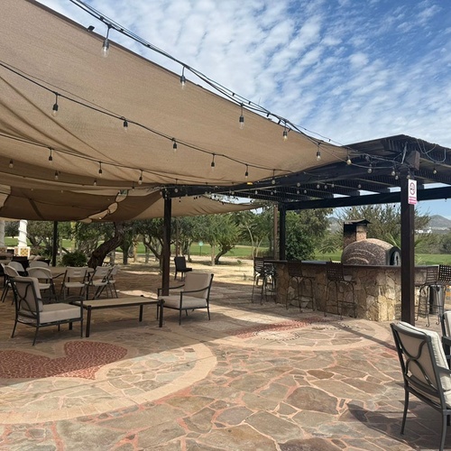 Un amplio patio exterior con toldos de tela y luces, un bar de piedra con horno de pizza, y diversas mesas y sillas, enmarcado por un campo de golf, montañas y cactus bajo un cielo nublado.