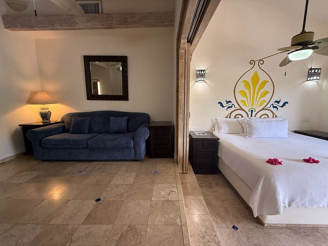 Una habitación espaciosa con suelo de baldosas muestra una zona de estar con sofá azul y una zona de dormitorio con cama blanca, separadas por una viga de madera.