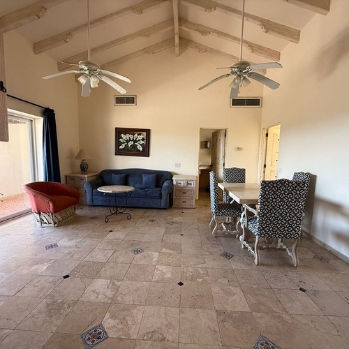 Una espaciosa sala-comedor de estilo rústico con techos altos de vigas de madera, dos ventiladores de techo, suelo de baldosas claras y mobiliario variado.