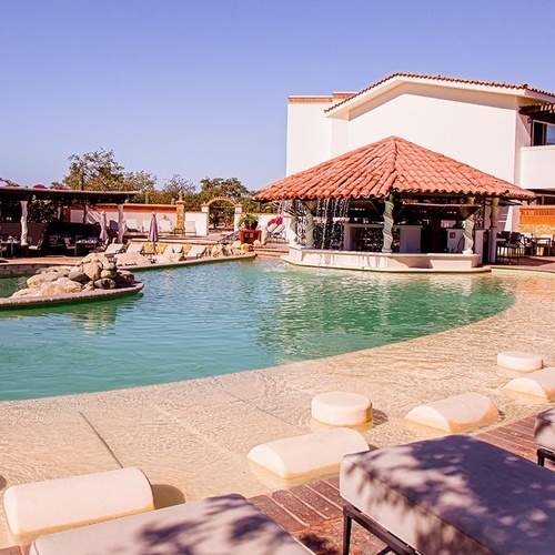 La imagen muestra una soleada piscina de hotel con un bar acuático de techo de tejas, rodeada de tumbonas y edificios de estilo mediterráneo.