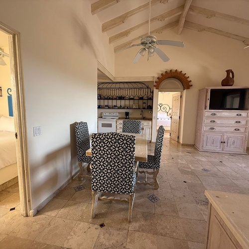 La imagen muestra el interior de una suite rústica con un dormitorio adyacente, área de comedor, cocina compacta y un mueble con televisor, bajo un techo abovedado con vigas de madera.