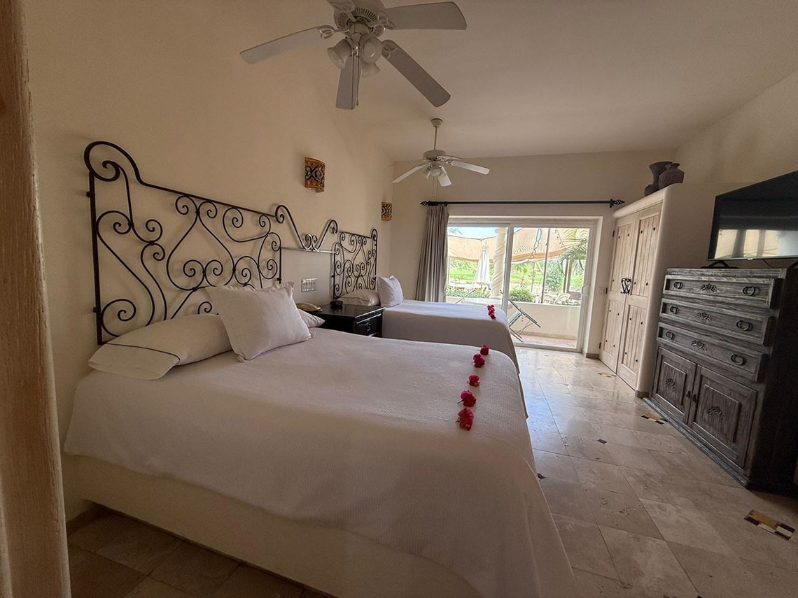 Una habitación luminosa de hotel presenta dos camas con cabeceros de hierro forjado ornamentados, y una puerta corredera que da a un patio exterior con vegetación.