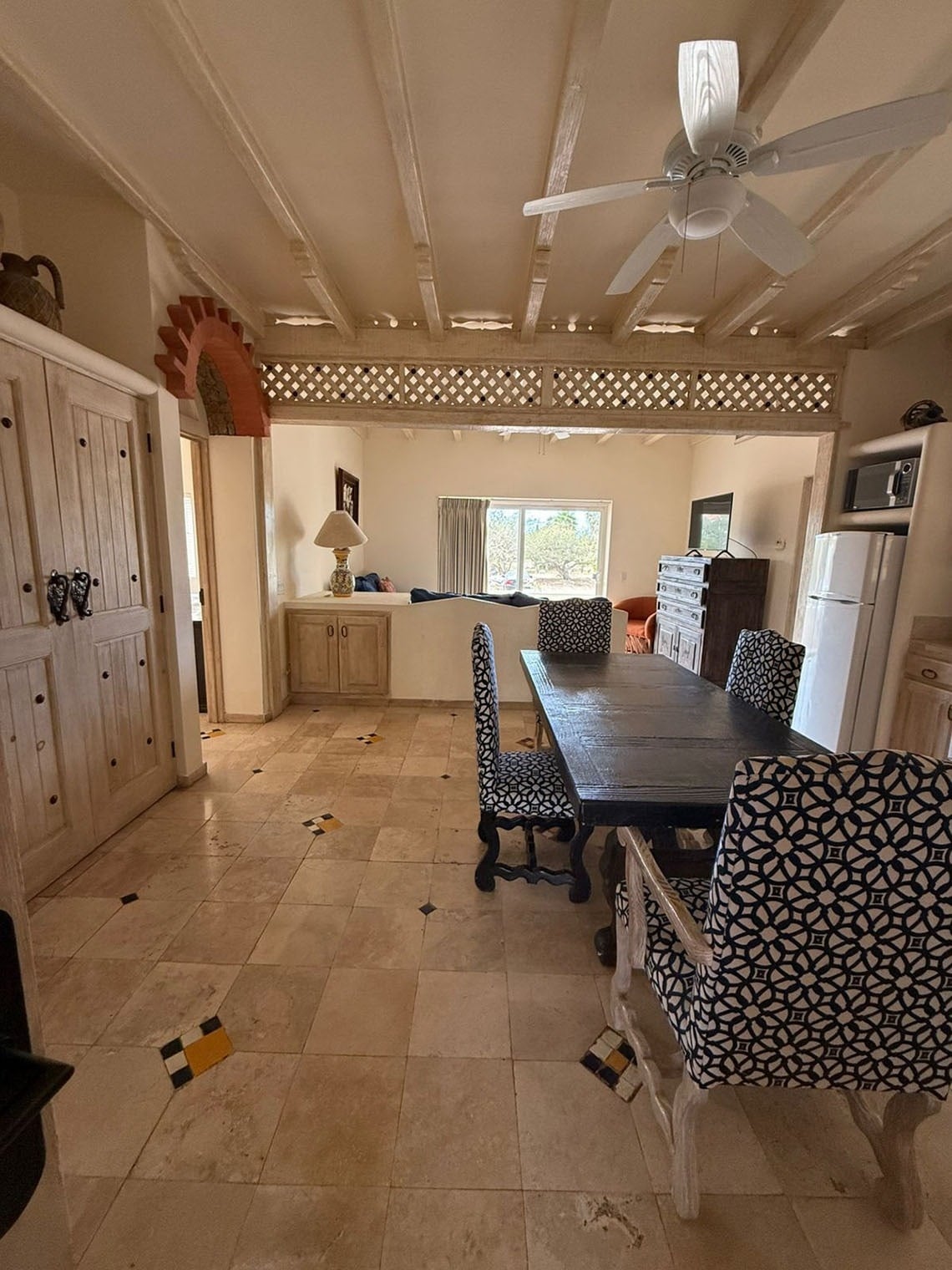 Un comedor de estilo rústico presenta una mesa de madera oscura con sillas tapizadas con patrón geométrico, sobre un suelo de baldosas claras con incrustaciones, y un techo con vigas de madera a la vista, conectado a otra estancia por un arco de ladrillo y una celosía de madera.