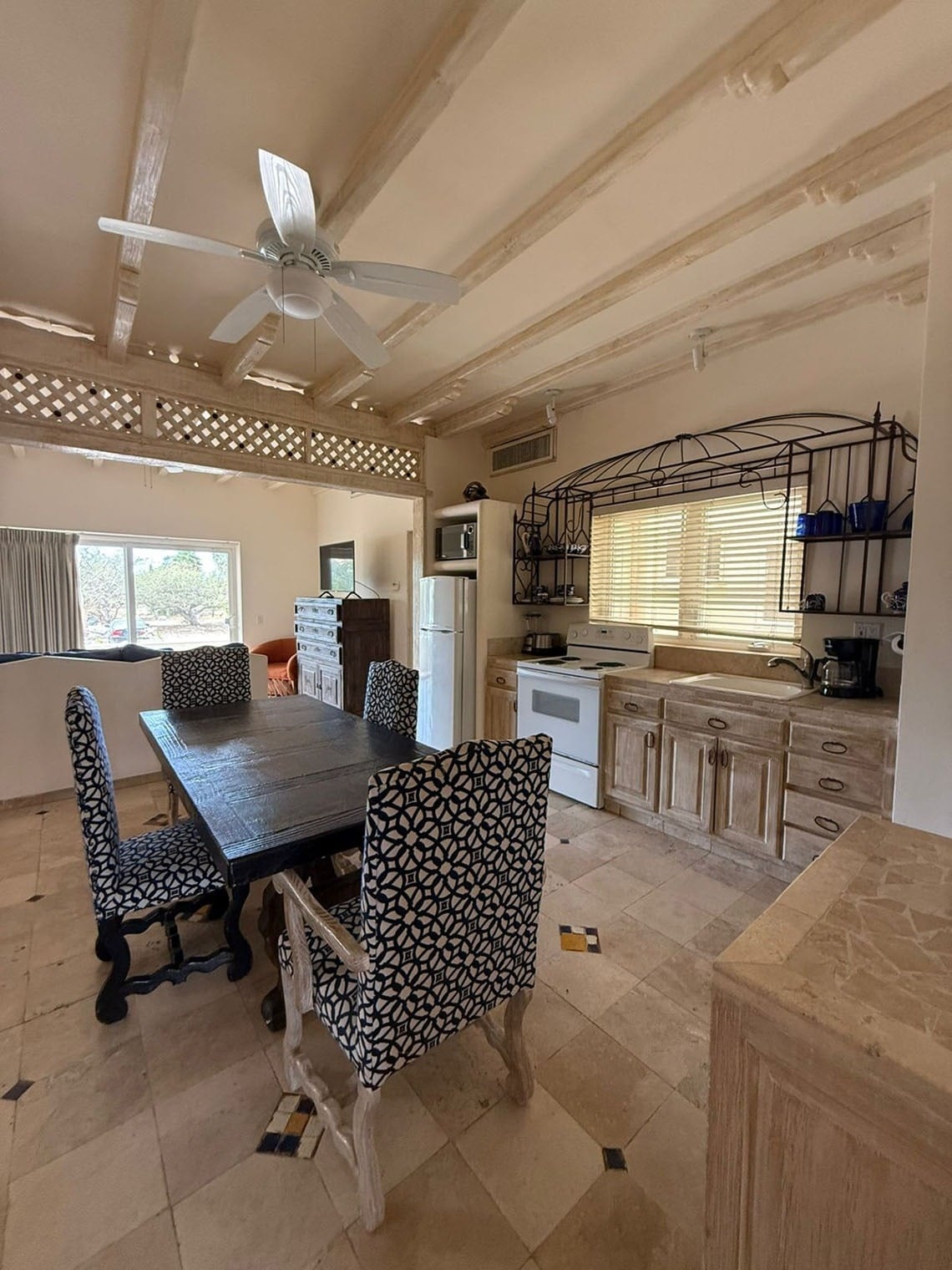 Una cocina y comedor de estilo rústico con vigas de madera claras en el techo, un ventilador de techo, una mesa de madera oscura rodeada de sillas tapizadas con un patrón geométrico, y un suelo de baldosas claras.