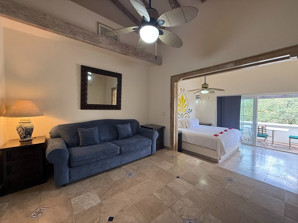 Una espaciosa suite presenta una sala de estar con un sofá azul y un dormitorio con una cama blanca, ambos bajo ventiladores de techo y con acceso a un balcón.