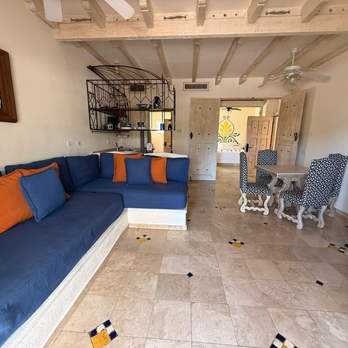 Una acogedora sala de estar y comedor de estilo rústico, con un gran sofá seccional azul y cojines naranjas, una mesa de comedor de madera clara con sillas estampadas, y un techo con vigas de madera, todo sobre un suelo de baldosas con detalles decorativos.