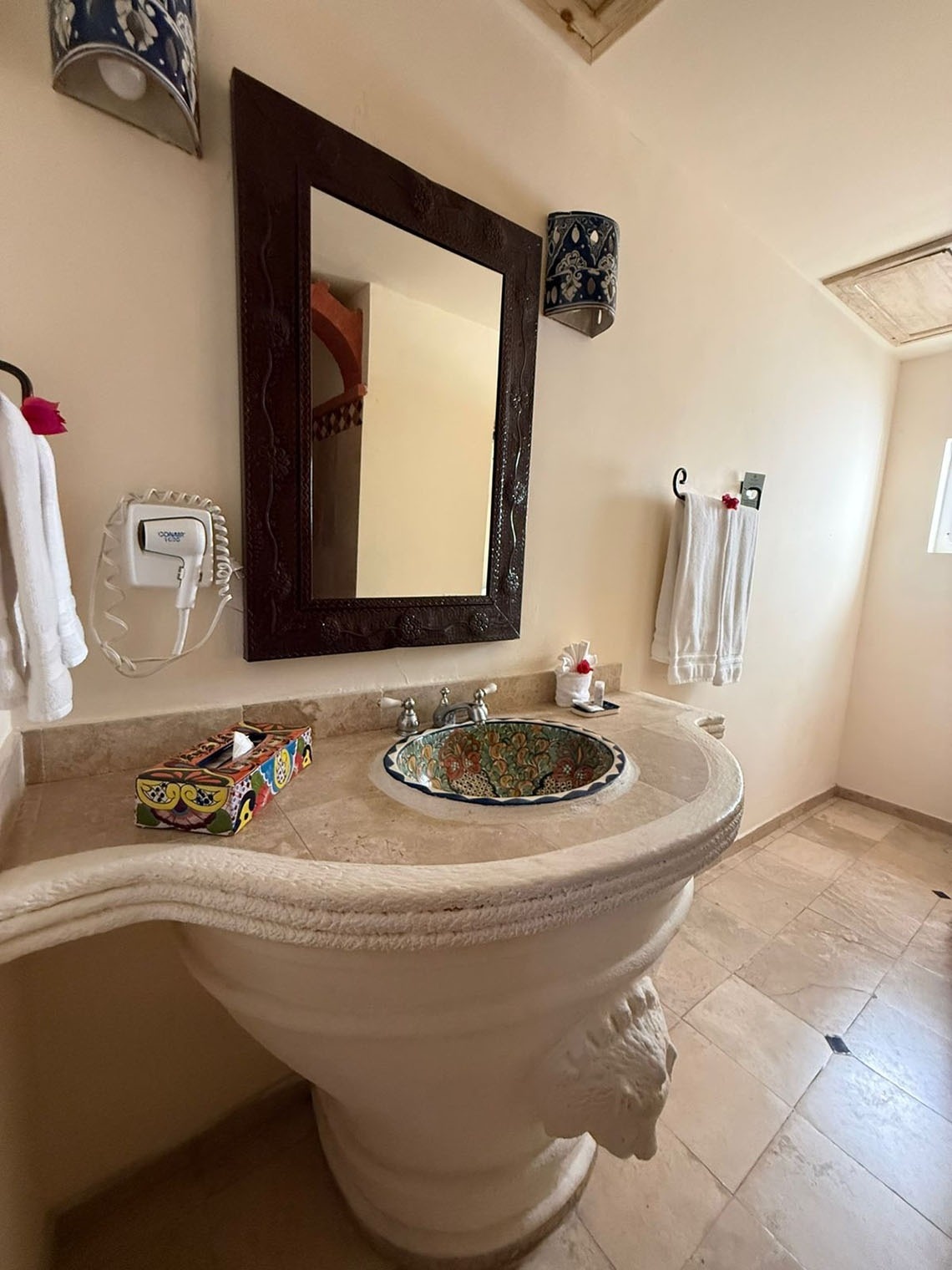 Un baño de estilo tradicional presenta un lavabo de pedestal tallado con una pileta de azulejos coloridos, un gran espejo de marco oscuro y toallas blancas colgadas en paredes claras.