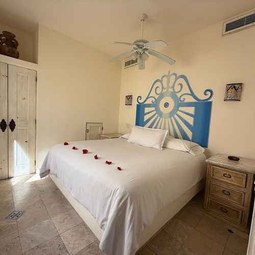 Una habitación luminosa y acogedora con una cama de matrimonio decorada con pétalos de rosa, un cabecero azul distintivo, suelo de baldosas, un armario de madera rústica y un ventilador de techo.