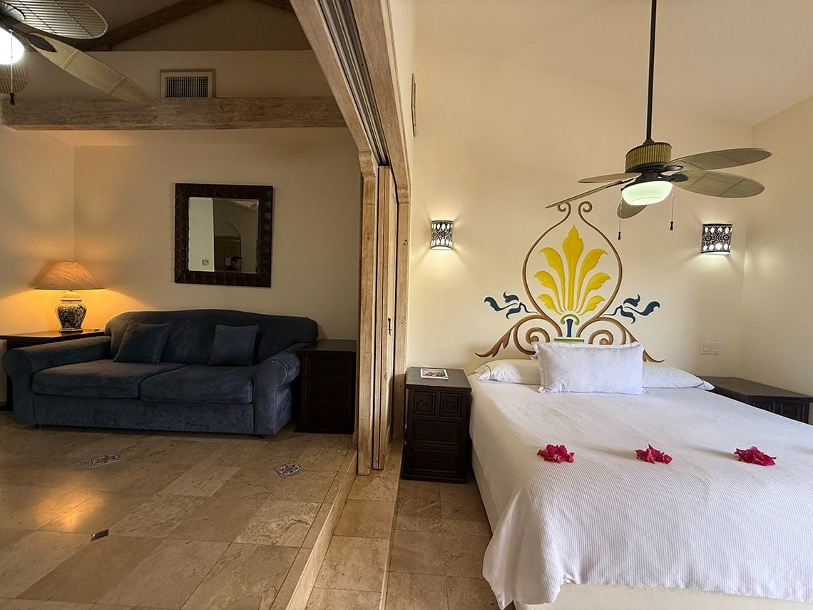 La imagen presenta una acogedora suite con un dormitorio decorado con motivos florales y un área de sala de estar con sofá, separadas por un nivel y equipadas con ventiladores de techo.