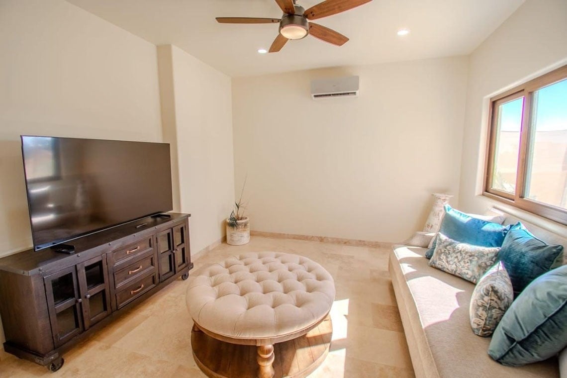 Una sala de estar moderna y luminosa con un televisor grande, un sofá con cojines azules, una otomana central y un ventilador de techo.