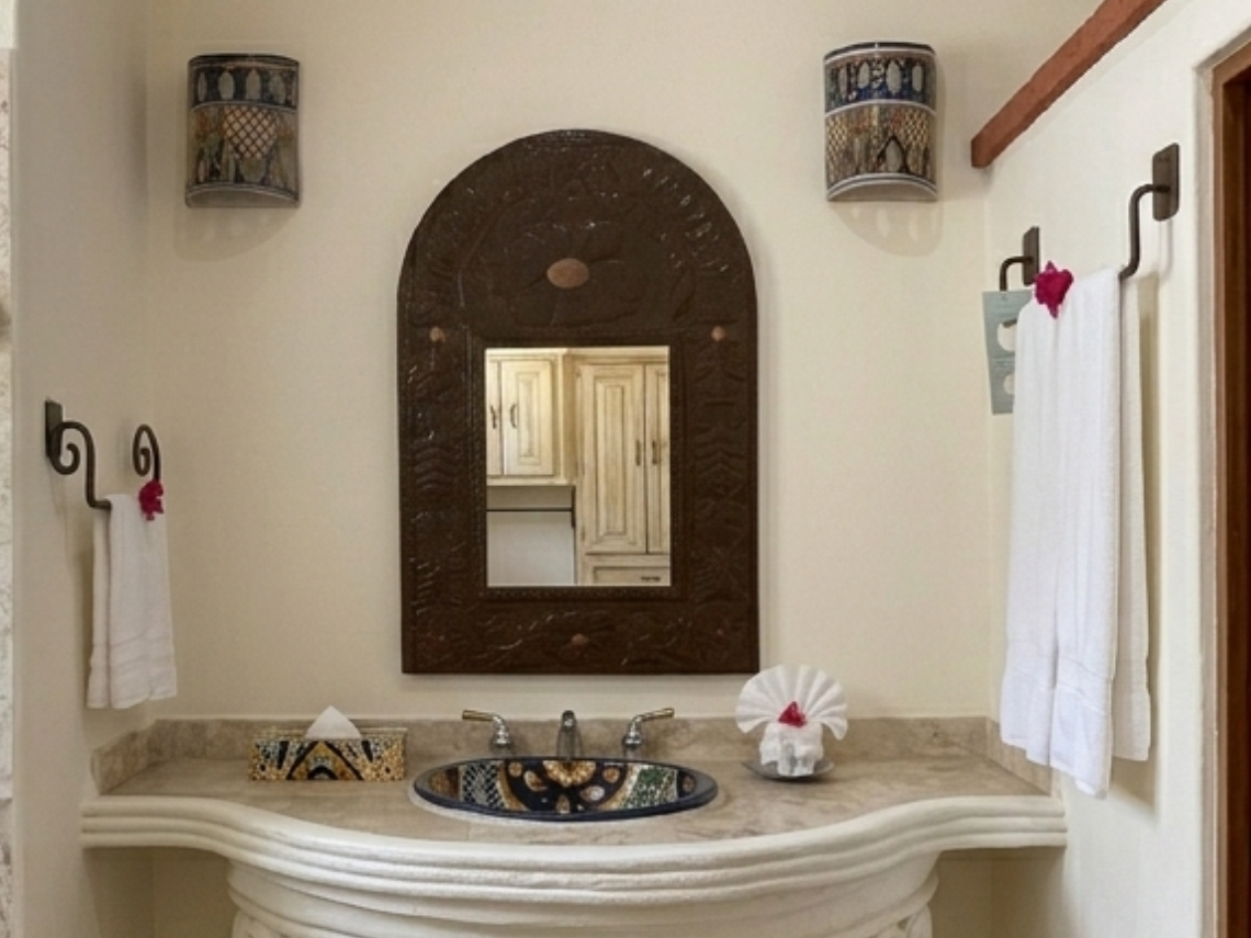 This image captures a bright bathroom featuring a tiled shower with a decorative border, separated by a rustic red-brown wall from a vanity area with a unique pedestal sink, ornate mirror, and a small window.