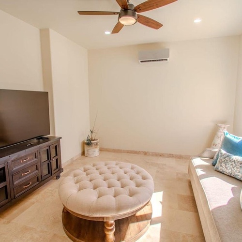 Una sala de estar bien iluminada presenta un televisor grande en un mueble oscuro, un otomán capitoné beige, un sofá con cojines azules, un ventilador de techo y una ventana.