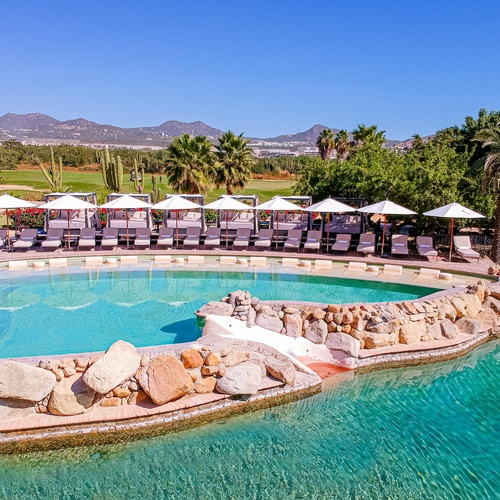 This aerial shot captures a sprawling Mediterranean-style resort complex featuring multiple pools, cabanas, and lounge areas, surrounded by lush vegetation with distant mountains in the background.