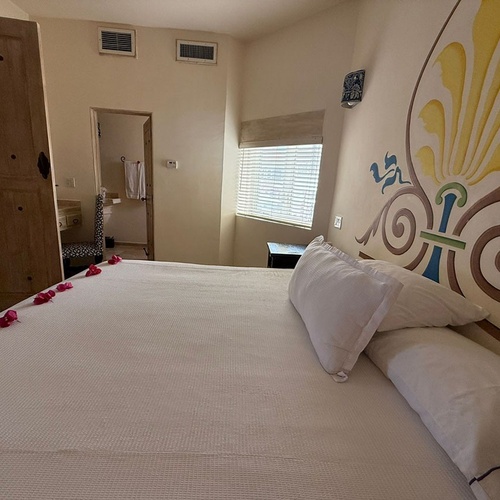 Un dormitorio con una cama blanca decorada con pétalos rosados, un gran mural artístico en la pared del cabecero y una entrada a un baño visible al fondo.