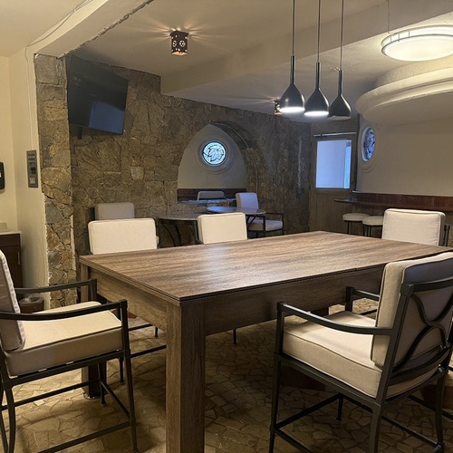 Una sala interior presenta una mesa de madera con sillas acolchadas, paredes de piedra decorativas, una barra con taburetes y diversas luminarias.
