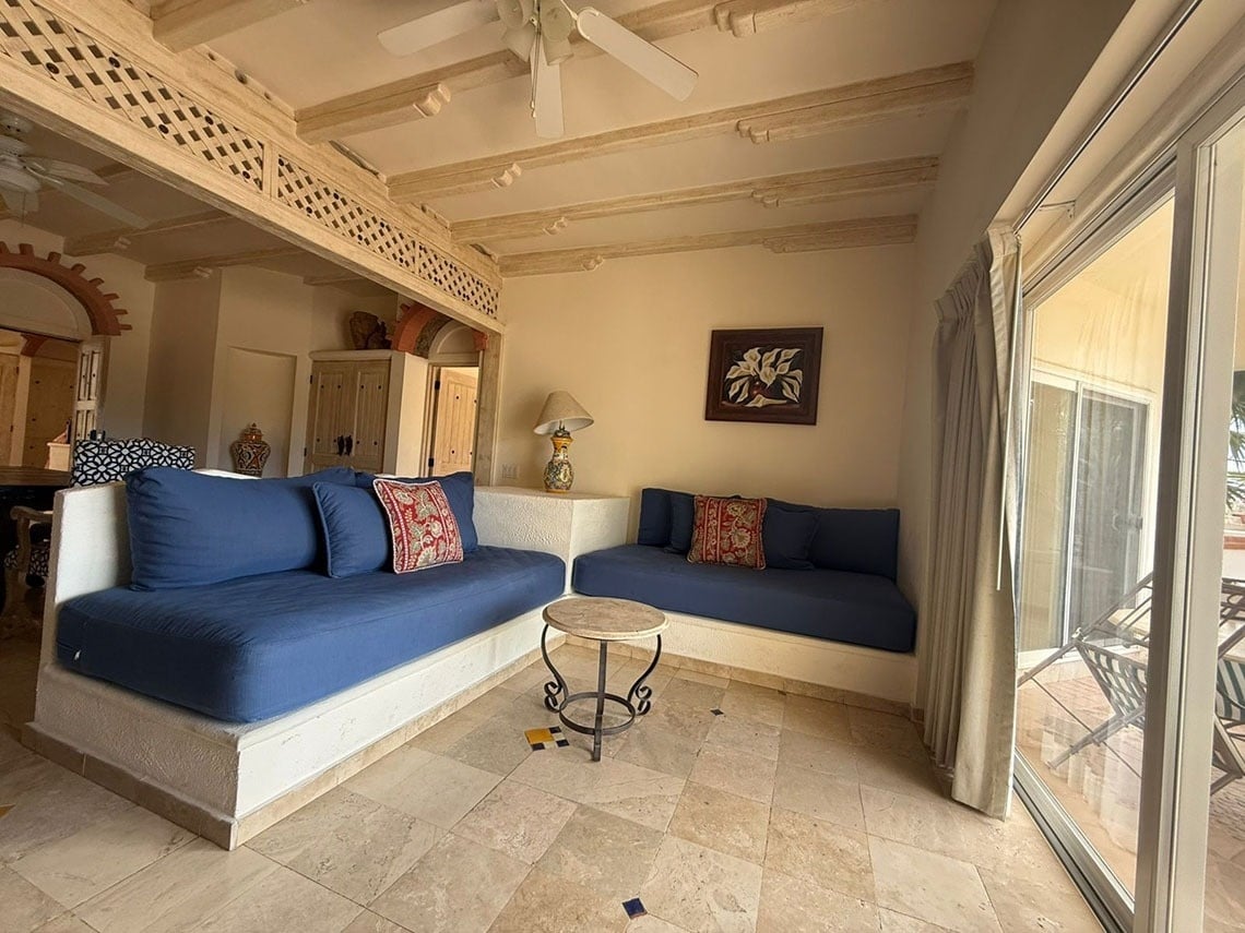 Una sala de estar de estilo mediterráneo con sofás azules empotrados en L, suelo de travertino y techo de vigas de madera, con acceso a un patio.