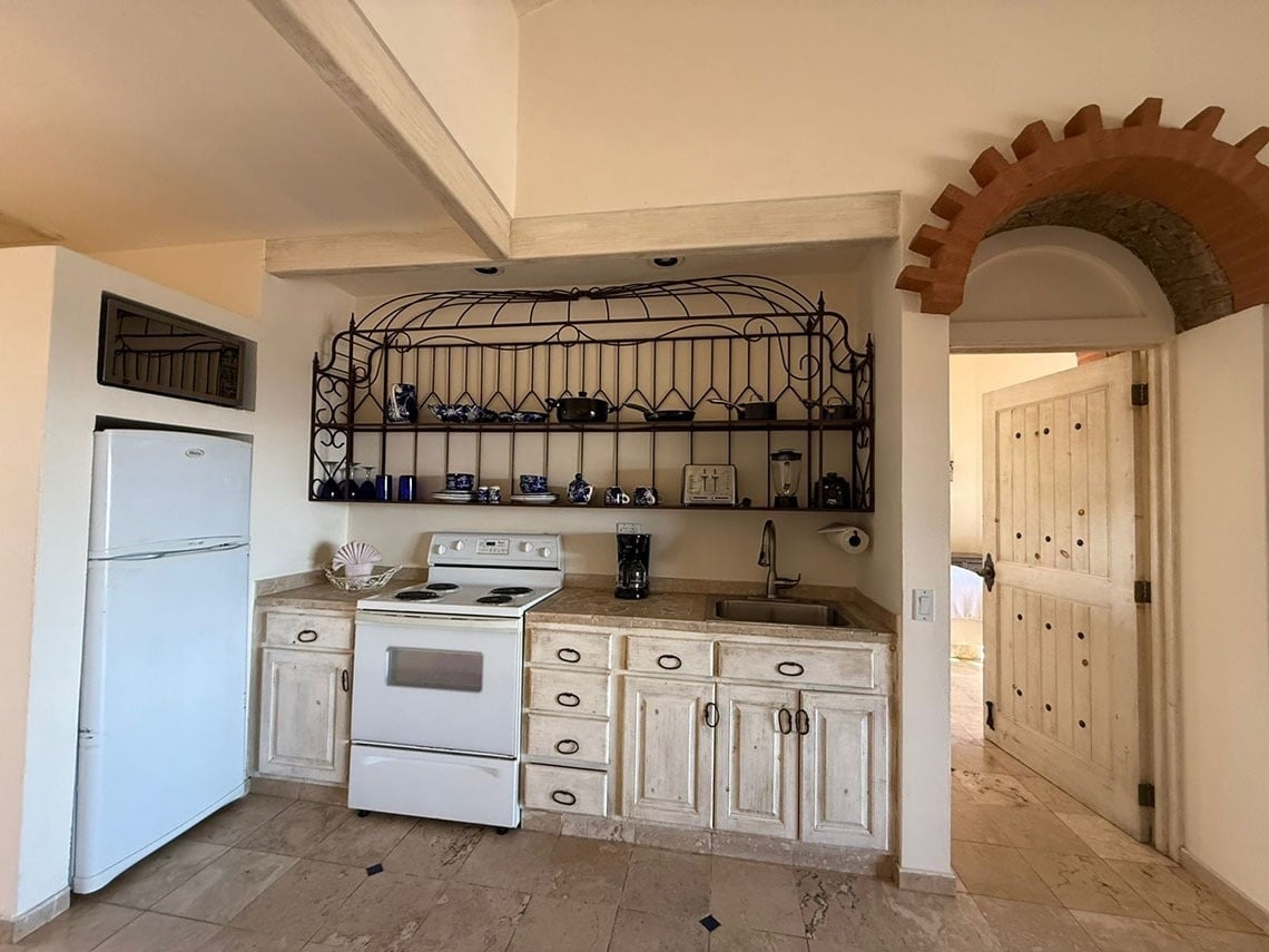 Una cocina de estilo rústico presenta gabinetes de madera clara, electrodomésticos blancos, una estantería de hierro forjado y una entrada con puerta de madera rústica bajo un arco de ladrillo decorativo.