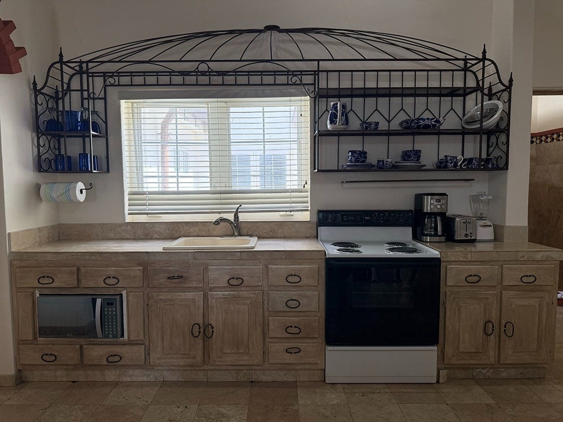 Una cocina de estilo rústico con un fregadero bajo una ventana, decorada con una gran estantería de hierro forjado sobre ella y gabinetes de madera.