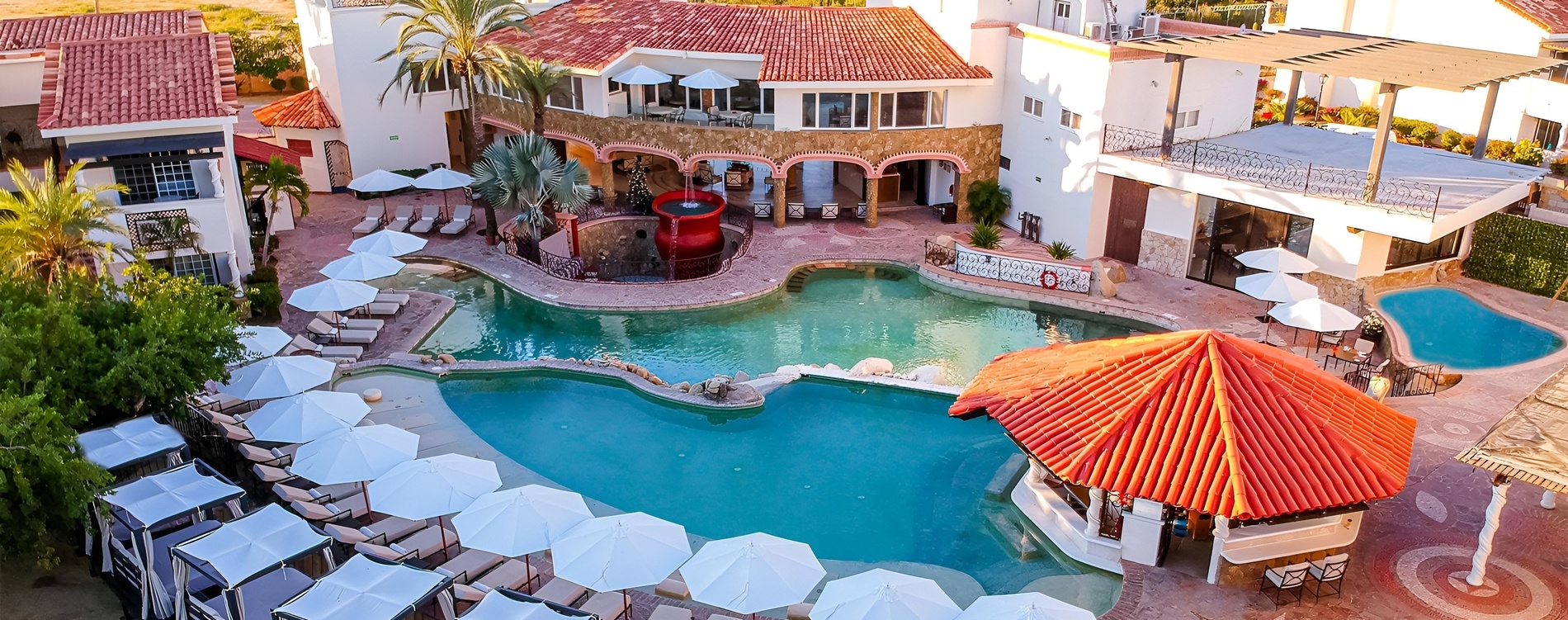 Un edificio de estilo mediterráneo o desértico, con paredes blancas y base de piedra, está flanqueado por grandes cactus bajo un cielo azul claro.