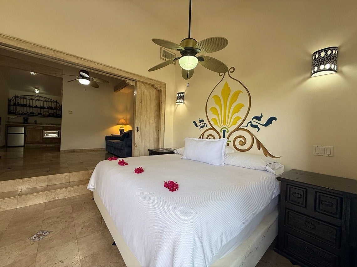 Un luminoso dormitorio con una cama blanca adornada con pétalos rojos y un vibrante mural en la cabecera, se conecta a un área de sala de estar o cocina a través de una entrada elevada.