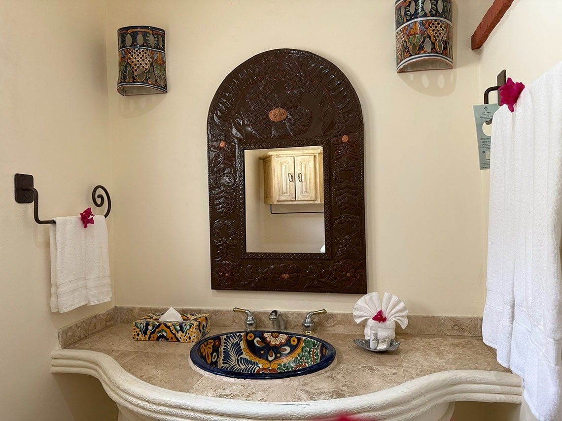 Un tocador de baño de estilo mexicano presenta un espejo de marco elaborado, un lavabo de cerámica pintado a mano y apliques decorativos en la pared.