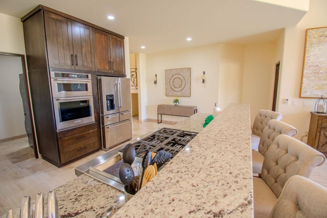 Una cocina moderna presenta gabinetes de madera oscura, electrodomésticos de acero inoxidable, y una isla de granito con una estufa integrada y asientos de barra.