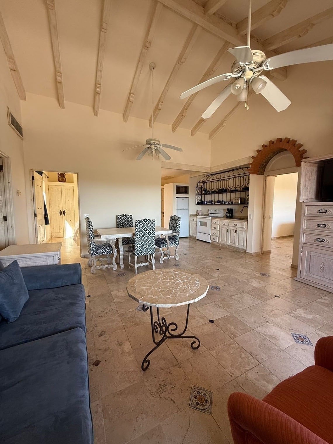 Amplia sala de estar de planta abierta que integra comedor y cocina, con techos altos de vigas expuestas, dos ventiladores de techo y suelos de baldosas con detalles decorativos.