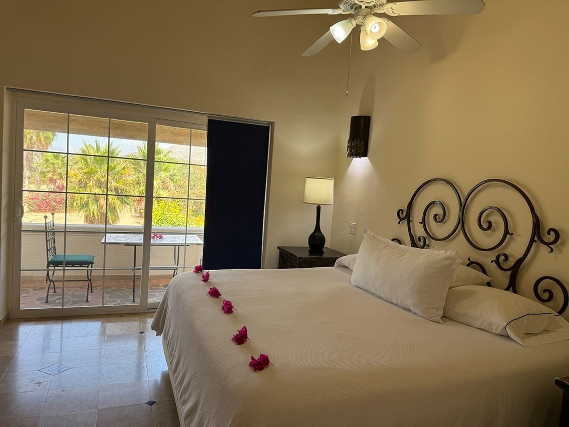 Una acogedora habitación de hotel cuenta con una cama grande decorada con flores rosadas, un ventilador de techo y un balcón con puertas correderas de cristal que dan a un paisaje de palmeras y plantas florecidas.